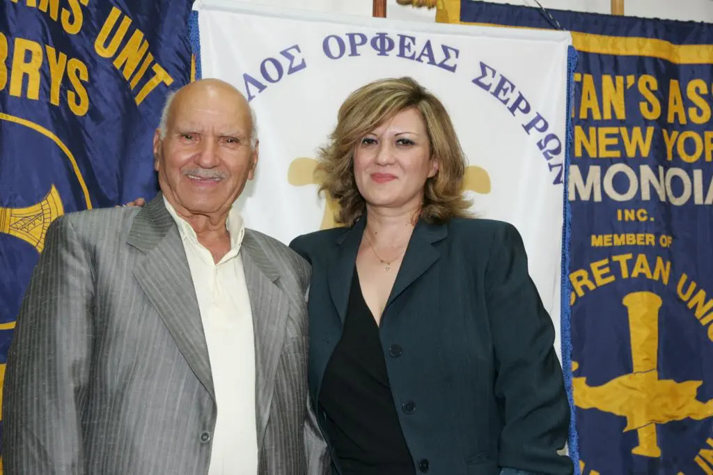 An elderly man and woman posing together indoors in front of a banner.