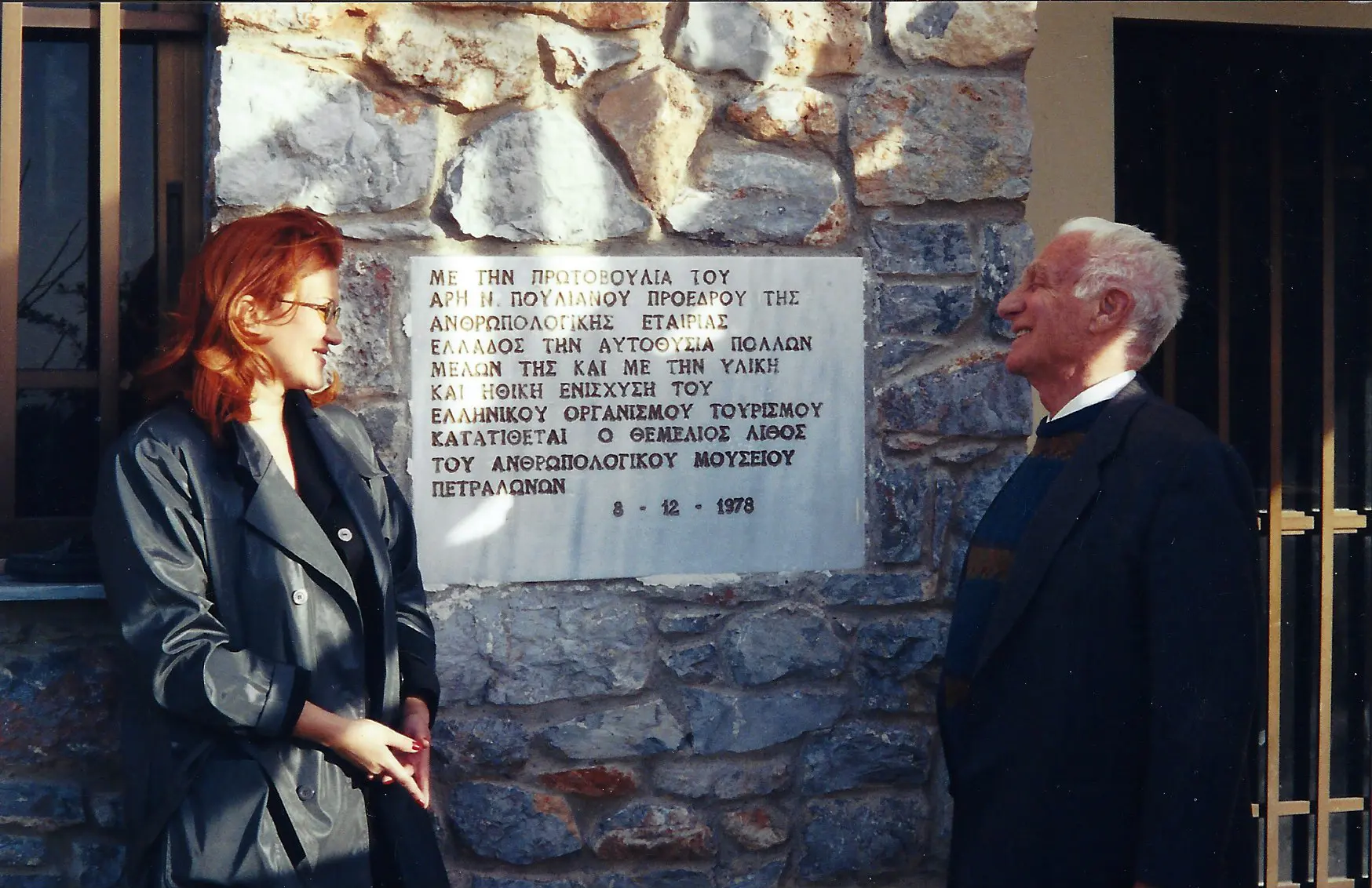 Two people stand near a commemorative plaque on a stone wall.
