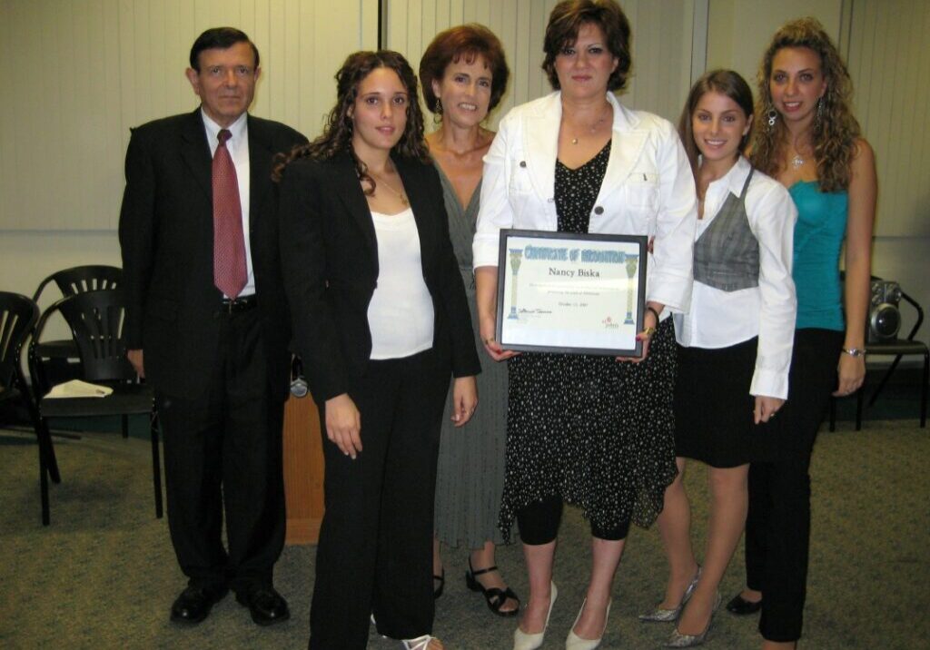 Five professionally dressed individuals pose indoors, one holding a certificate.