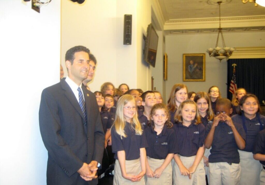 A man stands in front of a group of children dressed in uniforms in a formal room.